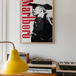 Op de afbeelding: Een ingelijste poster met een zwart-wit afbeelding van een rokende cowboy, met het rode woord "Marlboro" verticaal. Daaronder een houten kast met een gele lamp, audioapparatuur en een keyboard.