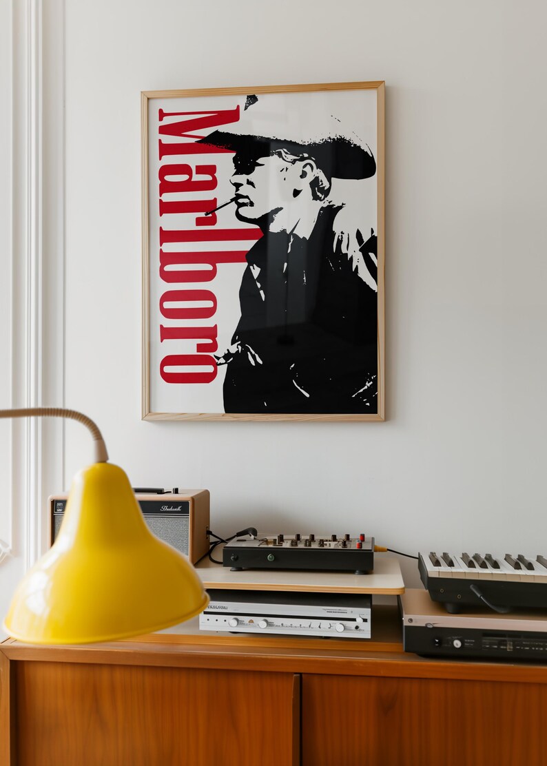Op de afbeelding: Een ingelijste poster met een zwart-wit afbeelding van een rokende cowboy, met het rode woord "Marlboro" verticaal. Daaronder een houten kast met een gele lamp, audioapparatuur en een keyboard.