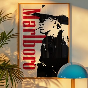 Op de afbeelding: Ingelijste kunstprint met een zwart-wit silhouet van een rokende persoon, met het woord "Marlboro" in het rood. Het kunstwerk zit in een gouden lijst, met een blauwe lamp en een plant op de voorgrond.