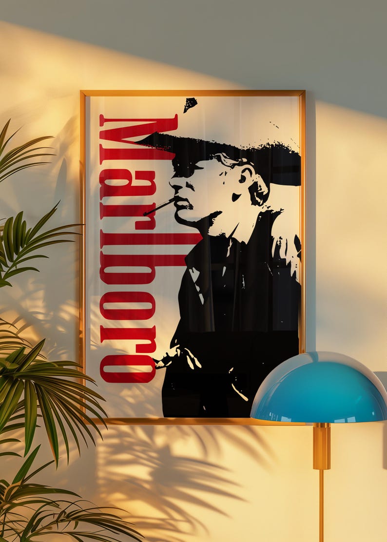 Op de afbeelding: Ingelijste kunstprint met een zwart-wit silhouet van een rokende persoon, met het woord "Marlboro" in het rood. Het kunstwerk zit in een gouden lijst, met een blauwe lamp en een plant op de voorgrond.