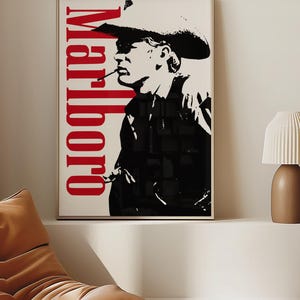 Op de afbeelding: Ingelijste kunstdruk met een zwart-wit afbeelding van een cowboy die een sigaret rookt. Het woord "Marlboro" is verticaal in het rood gedrukt. Het kunstwerk wordt gepresenteerd in een licht houten frame, geschikt voor wanddecoratie.