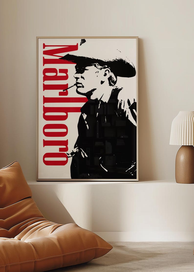 Op de afbeelding: Ingelijste kunstdruk met een zwart-wit afbeelding van een cowboy die een sigaret rookt. Het woord "Marlboro" is verticaal in het rood gedrukt. Het kunstwerk wordt gepresenteerd in een licht houten frame, geschikt voor wanddecoratie.