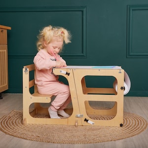 May include: A wooden learning tower for toddlers, featuring a chair and desk, with a paper roll attached. The child is wearing a pink outfit and is seated at the desk. The tower is made of natural wood.
