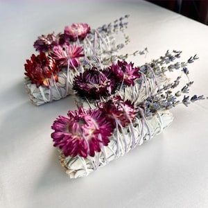 May include: Three white sage bundles adorned with dried purple flowers and lavender sprigs, tied with twine. The bundles feature vibrant red, pink, and purple blooms, creating a visually appealing arrangement. The sage bundles are arranged on a white surface.