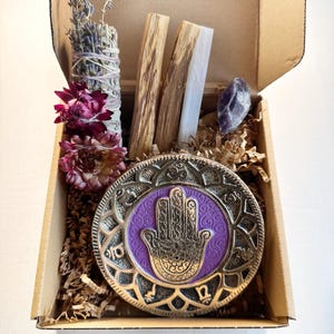 May include: A gift box filled with items for a spiritual practice. The box contains a purple and gold incense burner with a hamsa hand design, a bundle of sage, two sticks of palo santo wood, a clear crystal, and dried flowers.