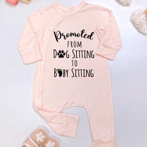 May include: A light pink baby onesie with long sleeves and snap closures. The text "Promoted from Dog Sitting to Baby Sitting" is printed in black. A pair of baby slippers and a rainbow decoration are also visible.