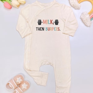 May include: Cream-colored baby onesie with long sleeves and a wrap-style closure. The onesie features the text "MILK, THEN BURPEES" with a barbell graphic. Pink and white slippers with star details are at the bottom.