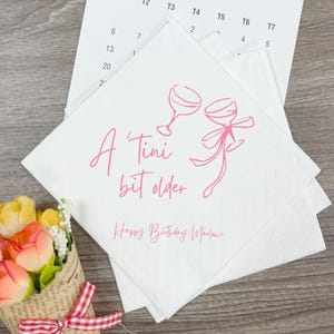 May include: White paper napkins with a pink design and text. The design includes two wine glasses and the phrase "A Tini bit older". The text "Happy Birthday Marlawe" is also present.