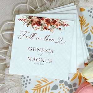 May include: White paper napkins with a floral design in shades of red, orange, and brown. The text "Fall in love" is written in a script font, with the names "Genesis and Magnus" and the date "10.06.2025" below.