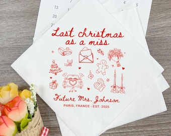 May include: White paper napkins with red text and illustrations. The text reads "Last christmas as a miss" and "Future Mrs. Johnson, PARIS, FRANCE - EST. 2025". The illustrations include a car, rings, and a Christmas tree.