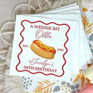 Könnte beinhalten: Quadratische weiße Servietten mit rotem Wellenrand. Die Servietten zeigen den Text "A Weenie Bit Older" mit einer Hotdog-Illustration und dem Text "Jocelyn's 30th Birthday". Das Jahr "1995" ist ebenfalls aufgedruckt.