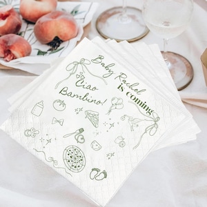 May include: White paper napkins with a green design and the text "Baby Rachel is coming" and "Ciao Bambino!". The napkins feature illustrations of baby-related items like a bottle, pizza, and footprints. Peaches and a glass of wine are in the background.
