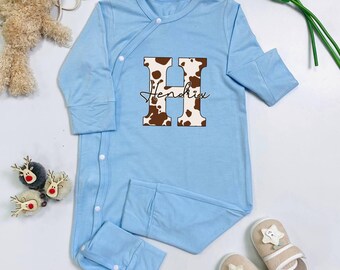 May include: A light blue baby sleeper with long sleeves and snap closures. The sleeper features a large letter "H" with a brown and white cow print design, and the name "Hendrix" in cursive script. A pair of beige slippers with star accents are also visible.