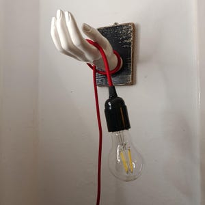 May include: A white hand sculpture holds a red cord, supporting a black light socket and a clear light bulb. The hand is mounted on a distressed, dark wooden plaque. The light fixture is attached to a wall.