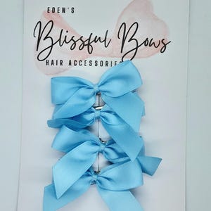 May include: A set of five light blue satin hair bows with silver clips. The bows are arranged in a stack on a white card with the text "Eden's Blissful Bows Hair Accessories".