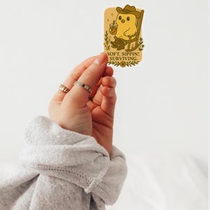 May include: A sticker showing a cartoon chick in a cowboy hat and boots, seated in a rocking chair and holding a drink. The sticker has the text "SOFT. SIPPIN': SURVIVING." in a serif font. The sticker is held by a hand.