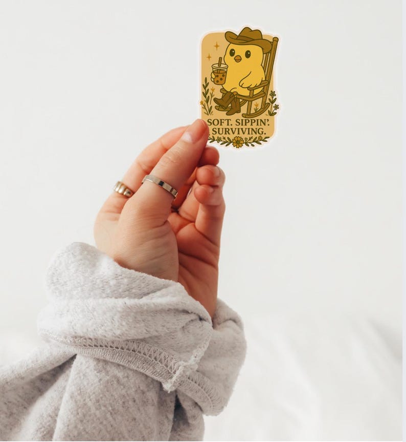 May include: A sticker showing a cartoon chick in a cowboy hat and boots, seated in a rocking chair and holding a drink. The sticker has the text "SOFT. SIPPIN': SURVIVING." in a serif font. The sticker is held by a hand.