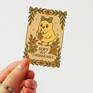 May include: A sticker with a cartoon yellow chick wearing a green bow, holding a drink with a straw. The sticker has a floral border and the text "SOFT BUT UNSHAKABLE".