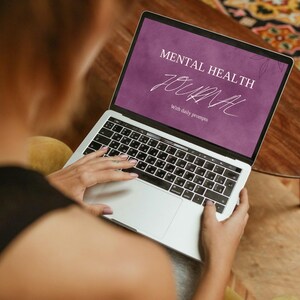 May include: A person using a silver laptop with a black keyboard. The screen displays the words "MENTAL HEALTH JOURNAL" in white text on a purple background, with the words "with daily prompts" below. The laptop is on a wooden table.