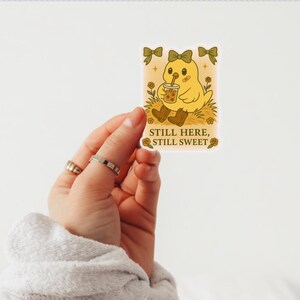 May include: A yellow sticker with a cartoon chick drinking bubble tea. The chick has a bow and is wearing brown boots. The sticker has the text "Still Here, Still Sweet". The background has decorative bows.