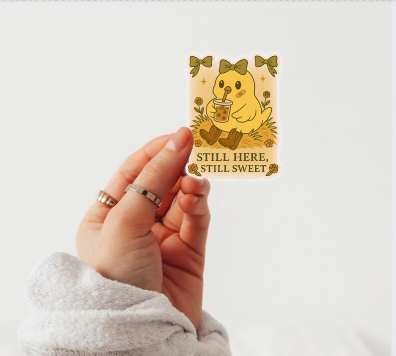 May include: A yellow sticker with a cartoon chick drinking bubble tea. The chick has a bow and is wearing brown boots. The sticker has the text "Still Here, Still Sweet". The background has decorative bows.