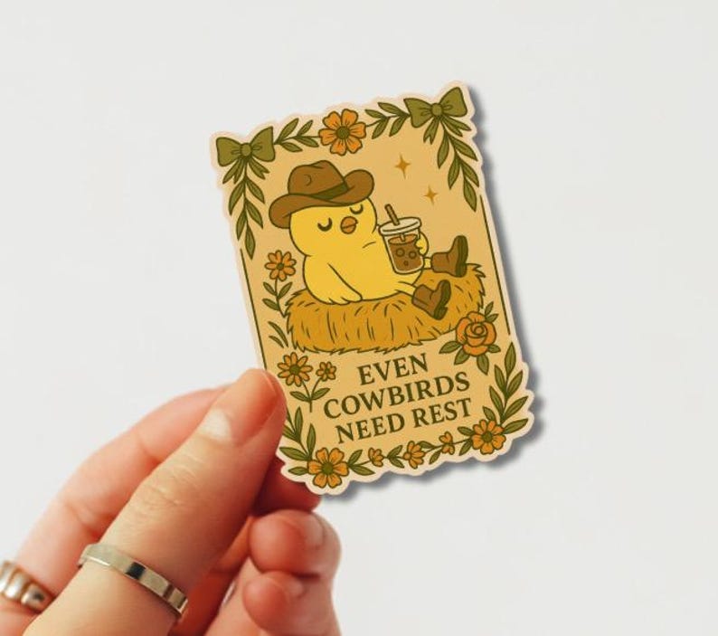 May include: A sticker with a yellow cartoon cowbird in a cowboy hat and boots, holding a drink. The cowbird is resting on hay. The sticker has a floral border and the text "EVEN COWBIRDS NEED REST".
