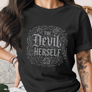 Puede incluir: Camiseta negra con la frase "The Devil Herself" en una fuente gótica, rodeada de un diseño floral y espinoso. El diseño es en tonos grises y blancos, creando un contraste llamativo sobre la tela oscura.