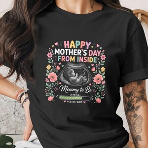 Può includere: T-shirt nera con un design grafico colorato per la festa della mamma. Il design include il testo "Happy Mother's Day From Inside" e un'immagine ecografica di un bambino, circondata da fiori.