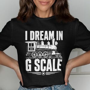 Può includere: T-shirt nera con grafica bianca di una locomotiva a vapore e la scritta "I DREAM IN G SCALE". L'illustrazione del treno include il numero "48". Il disegno è centrato sulla maglietta.