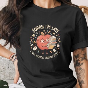 May include: Black t-shirt featuring a cartoon apple character wearing glasses and holding books. The text on the shirt reads "SORRY I'M LATE I WAS AVOIDING GRADING PAPERS". The design includes stars, pencils, and a coffee cup.