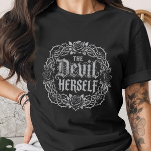 Puede incluir: Camiseta negra con un diseño gótico. El diseño presenta el texto "THE Devil HERSELF" dentro de un círculo de rosas y espinas. La camiseta tiene mangas cortas.