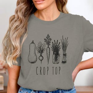 May include: A grey crop top with a black and white illustration of vegetables, such as a squash, radish, carrot, and herbs. The words "CROP TOP" are printed below the illustration in a simple font.