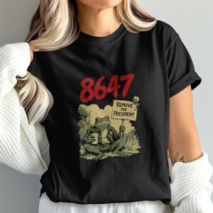May include: Black t-shirt with a graphic of a frog holding a sign that says "Remove the President." The number "8647" is printed above the frog in red. The frog is wearing a jacket and holding a cup.