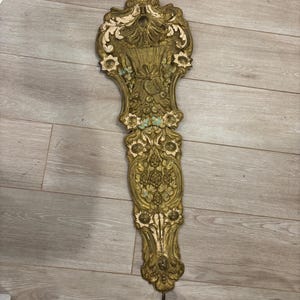 May include: Ornate, antique wall decor in gold and off-white. The piece features a detailed, symmetrical design with floral and scrollwork motifs. A metal hook extends from the top, and a metal rod hangs from the bottom.