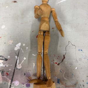 May include: A wooden artist's mannequin with articulated joints, standing on a small wooden base. The mannequin is made of light-colored wood and is designed to mimic the human form for drawing and sketching.