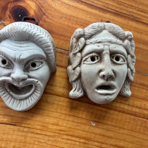 May include: Two off-white, theatrical masks depicting comedy and tragedy. The comedy mask has a wide, open mouth and furrowed brow. The tragedy mask has a sorrowful expression. Both masks have a loop for hanging.