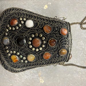 May include: A silver-toned, rectangular purse with a decorative filigree design. The purse is adorned with various colored, polished stones. A delicate chain strap is attached. The purse is likely vintage or antique.
