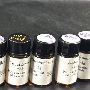 May include: A collection of small amber glass bottles with black caps, each labeled with handwritten text. The bottles contain pure essential oil blends, with labels indicating the scent and weight, such as "Oud Cohban" and "Floria."