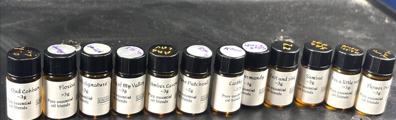 May include: A collection of small amber glass bottles with black caps, each labeled with handwritten text. The bottles contain pure essential oil blends, with labels indicating the scent and weight, such as "Oud Cohban" and "Floria."