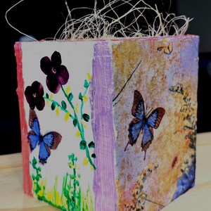 May include: A decorative cube with painted floral and butterfly designs. The cube features a white background with purple flowers and a blue butterfly. The sides have a textured, multi-colored design with additional butterflies and floral elements.