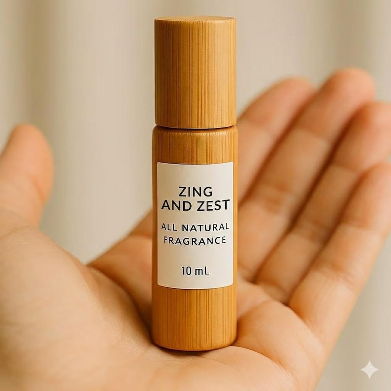 May include: A small, cylindrical fragrance bottle with a wooden cap and body is held in an open hand. The label reads "ZING AND ZEST ALL NATURAL FRAGRANCE" and "10 mL". The bottle has a natural, light brown color.