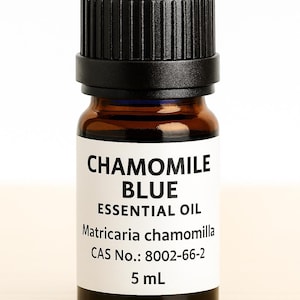 May include: A small amber glass bottle of Chamomile Blue Essential Oil with a black cap. The label displays the product name, botanical name, and volume of 5 mL. The bottle is filled with a blue liquid.