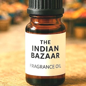 May include: A small, brown glass bottle of fragrance oil with a black cap. The label reads "THE INDIAN BAZAAR FRAGRANCE OIL" in bold, black letters. The bottle sits on a rough, wooden surface, with a blurred background.