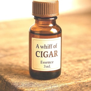 May include: A small, brown glass bottle with a brown cap. The label reads "A whiff of CIGAR Essence 3 mL". The bottle is sitting on a wooden surface. The bottle is filled with a dark liquid.