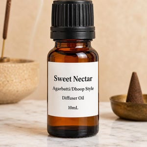 May include: A brown glass bottle with a black cap, labeled "Sweet Nectar Agarbattti/Dhoop Style Diffuser Oil 10mL." Incense and a cone-shaped object are in the background. The bottle is filled with a dark amber liquid.