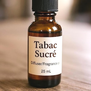 May include: A small, brown glass bottle with a black cap. The label reads "Tabac Sucré, Diffuser/Fragrance Oil, 25 mL." The bottle is filled with a light brown liquid. The bottle is sitting on a wooden surface.