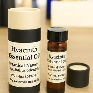 May include: Two containers of Hyacinth Essential Oil. One is a tall, cylindrical tube with a beige top and a black base. The other is a small, brown glass bottle with a black cap. Both have labels with product information.
