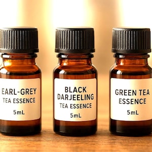 May include: Three small amber glass bottles with black caps and white labels. The labels read "EARL-GREY TEA ESSENCE 5mL", "BLACK DARJEELING TEA ESSENCE 5mL", and "GREEN TEA ESSENCE 5mL". The bottles are arranged in a row.