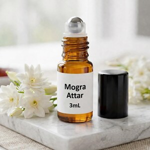 May include: A small amber glass roll-on bottle with a silver rollerball and a black cap. The label reads "Mogra Attar 3mL". White jasmine flowers are arranged on a marble surface next to the bottle.