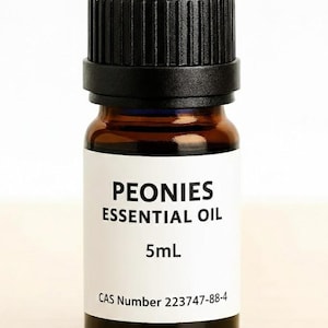 May include: A small amber glass bottle with a black cap. The label reads "PEONIES ESSENTIAL OIL" and "5mL". The CAS number 223747-88-4 is printed at the bottom of the label. The bottle is on a light wood surface.